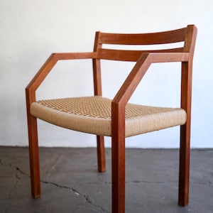 May include: A mid-century modern armchair with a woven seat and armrests. The chair features a solid wood frame in a warm brown tone. The seat is made of woven natural fibers. The chair has a minimalist design.