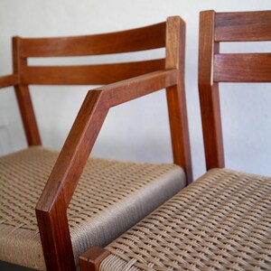May include: Two wooden chairs with woven seats. The chairs have a warm, brown finish and feature armrests. The seats are made of a light tan woven material, creating a contrast with the darker wood.