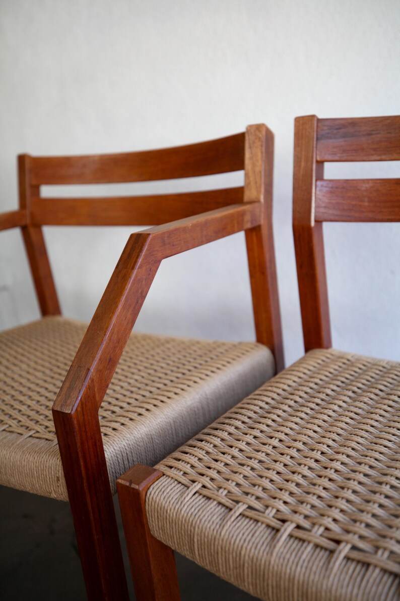May include: Two wooden chairs with woven seats. The chairs have a warm, brown finish and feature armrests. The seats are made of a light tan woven material, creating a contrast with the darker wood.