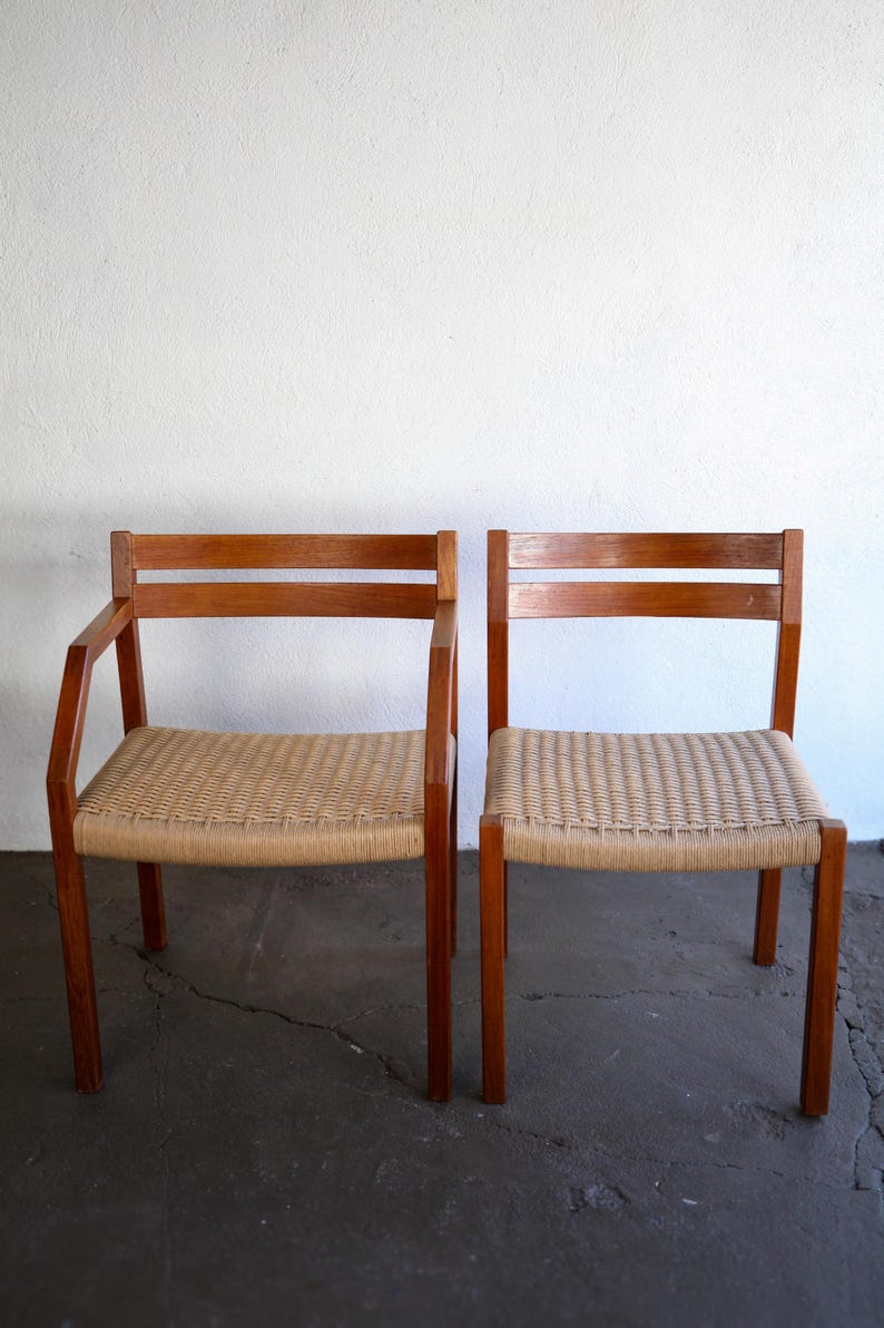 May include: Two wooden chairs with woven seats. The chairs have a light brown finish and a rectangular back with two horizontal slats. One chair has armrests. The seats are made of a light tan woven material.