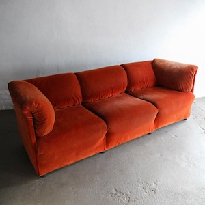 May include: A three-seat, burnt orange velvet sofa. The sofa has a low profile with a rounded arm on the left side and a square arm on the right. The cushions are square and the sofa sits on a dark base. The background is a plain white wall.