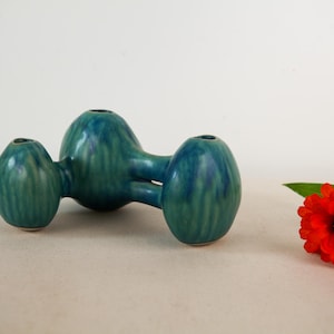 May include: A teal ceramic vase with three rounded, bulbous chambers connected by a central bridge, featuring a glossy, variegated glaze with darker blue streaks. A single bright red zinnia flower with green leaves sits to the right.