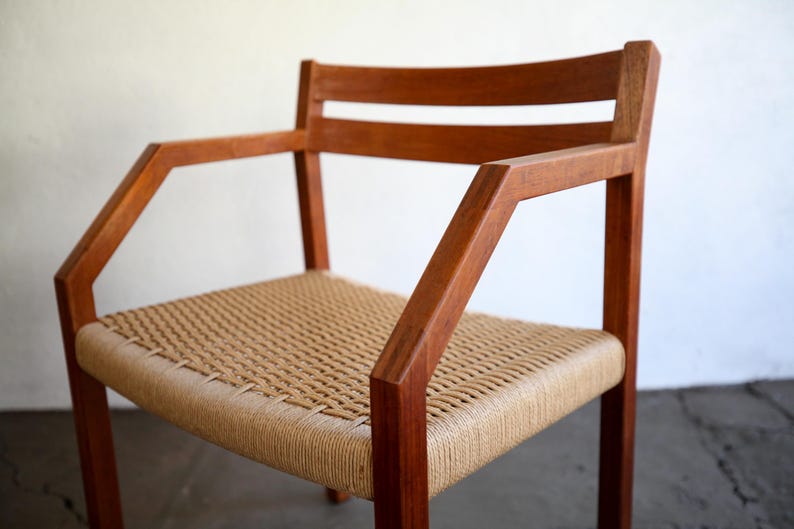 May include: A wooden armchair with a woven seat and back. The chair has a natural wood finish and angled armrests. The seat is made of a light tan woven material. The chair is a mid-century modern style.
