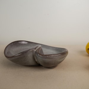 May include: A dual-compartment ceramic dish with a smooth, glossy finish in a muted mauve-gray tone. The bowl features a curved, organic shape with a central divider separating two distinct sections, creating a functional piece for serving.
