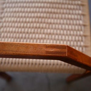 May include: Close-up of a wooden chair armrest with a woven seat. The armrest is made of reddish-brown wood, with visible joinery at the corner. The seat has a light beige woven pattern.