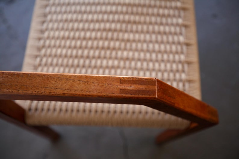 May include: Close-up of a wooden chair armrest with a woven seat. The armrest is made of reddish-brown wood, with visible joinery at the corner. The seat has a light beige woven pattern.
