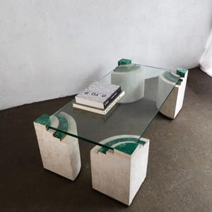 May include: A modern glass coffee table with a rectangular top. The table has four geometric, white stone legs with green accents. Two books are on the table. The table is a unique piece of furniture.