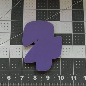 May include: Purple plastic cookie cutter shaped like the number 2. The cutter is on a black and white grid background.
