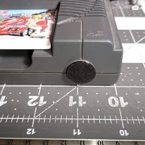 May include: Close-up of a gray video game console with a game cartridge inserted. The cartridge has a colorful image of a car and a woman. A black, round, felt pad is on the bottom of the console.