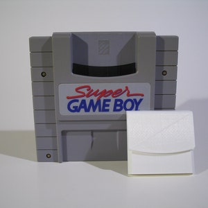 May include: A grey Super Game Boy cartridge with a white plastic case. The cartridge has a black slot for inserting a Game Boy game.