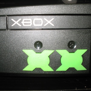 May include: A black Xbox console with the word "XBOX" printed on the front. The console has two green controller ports labelled "1" and "2".