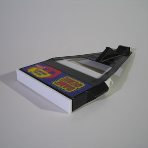 May include: Black plastic Game Genie cartridge holder with a white base. The holder has a blue and yellow label that says "Game Genie" and "Enhance Your Game".