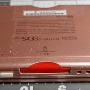 May include: Rose gold Nintendo DS Lite console. The back of the handheld gaming device features the Nintendo logo, model information, and a red game card slot. The text "NINTENDO DS lite" is printed on the device.