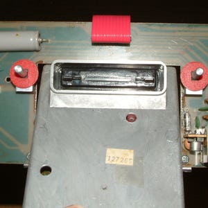 May include: A close-up of a circuit board with a silver metal component and red toggle switches. The circuit board has a green background and the component has the number "127367" printed on it.