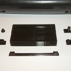 May include: A set of black plastic pieces for a video game console. The pieces include a rectangular base, a smaller rectangular piece, a long rectangular piece, and two small round pieces.