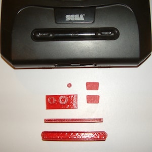 Peut inclure: Gros plan d'une console Sega Genesis noire avec un bouton rouge et un interrupteur d'alimentation rouge. La console est posée sur une surface blanche. Il y a plusieurs pièces en plastique rouge à côté de la console.