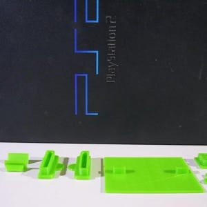 May include: A black PlayStation 2 console with the PlayStation 2 logo in blue. Several bright green 3D-printed components are arranged in front of the console, including a base and various support pieces.