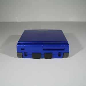 May include: A blue Game Boy Advance SP handheld gaming console. The console is closed and the screen is facing away from the viewer.
