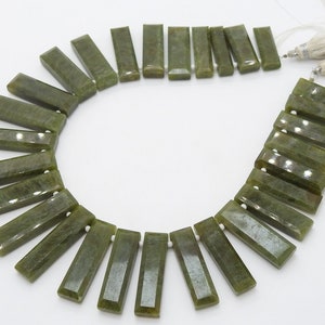 May include: A strand of olive green rectangular gemstone beads. Each bead is faceted and has a polished surface. The beads are strung together with white string and tassels. The gemstones are approximately 1 inch in length.