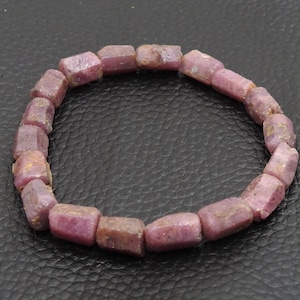 Può includere: Un braccialetto composto da pietre preziose rettangolari rosa-rossastre tagliate in modo grezzo. Il braccialetto è circolare e poggia su una superficie scura e strutturata. Le pietre hanno un aspetto naturale e non lucidato.
