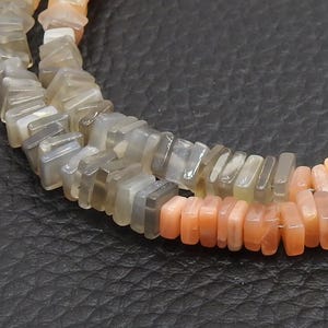 Puede incluir: Primer plano de dos hilos de cuentas de piedras preciosas. Las cuentas son rectangulares y varían en color de gris a melocotón. Las cuentas están ensartadas, creando un collar o pulsera.