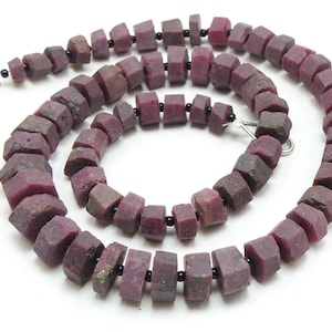 May include: A necklace of faceted, deep red ruby beads. The beads are irregularly shaped and strung together with black spacers. The necklace is displayed against a white background, highlighting the color and texture of the gemstones.