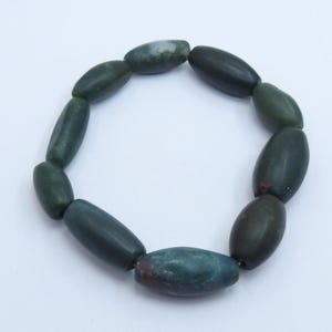 Puede incluir: Un brazalete de cuentas verde oscuro con cuentas de forma ovalada. Las cuentas tienen un acabado pulido y varían ligeramente en color, con algunos toques de rojo y verde más claro. El brazalete se muestra sobre una superficie blanca.