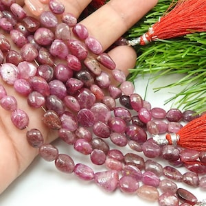 May include: A strand of polished, irregular, deep pink ruby gemstone beads with a smooth, glossy finish. The beads are strung together and accented with two vibrant red decorative tassels featuring silver-toned metal caps at the top ends.