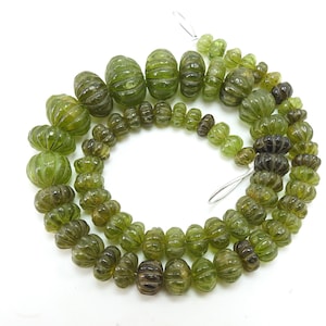 以下が含まれることがあります： さまざまなサイズと形状の緑色の宝石ビーズの連なり。リブや丸みを帯びたデザインが含まれています。ビーズは紐でつながれており、オリーブグリーンや濃いグリーンなど、さまざまな色合いが表現されています。白い背景にビーズが表示されています。