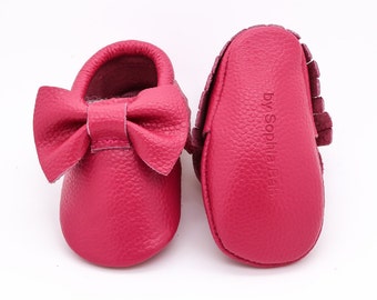May include: A pair of pink leather baby moccasins with a bow on the front. The moccasins have a fringe detail and the brand name "by Sophia Baby" is printed on the back.