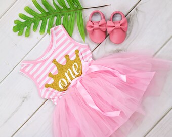May include: Pink and white striped baby dress with a gold glitter crown and the word "one" on the front. The dress has a tulle skirt and comes with a pair of pink baby shoes.