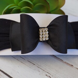 May include: A black baby headband with a large bow and a rhinestone embellishment. The bow is centered on a stretchy black band. The headband is displayed on a white card, with a green plant in the background.