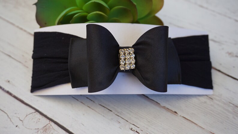 May include: A black baby headband with a large bow and a rhinestone embellishment. The bow is centered on a stretchy black band. The headband is displayed on a white card, with a green plant in the background.