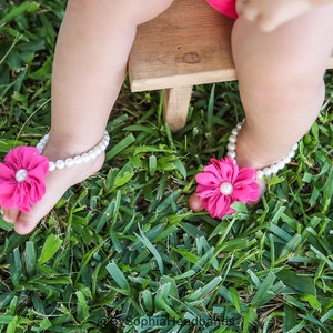 Pode incluir: Um par de sand&aacute;lias de flores rosa para p&eacute;s descal&ccedil;os com detalhes de p&eacute;rolas brancas. As sand&aacute;lias s&atilde;o usadas por um beb&ecirc; em uma superf&iacute;cie gramada.