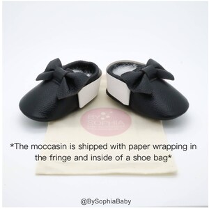 May include: A pair of black leather baby moccasins with a white trim and a black bow on each shoe. The moccasins are on a white and brown surface with the text "BY SOPHIA Baby Boutique" and "*The moccasin is shipped with paper wrapping in the fringe and inside of a shoe bag*" printed on the surface. The text "@BySophiaBaby" is also printed on the surface.