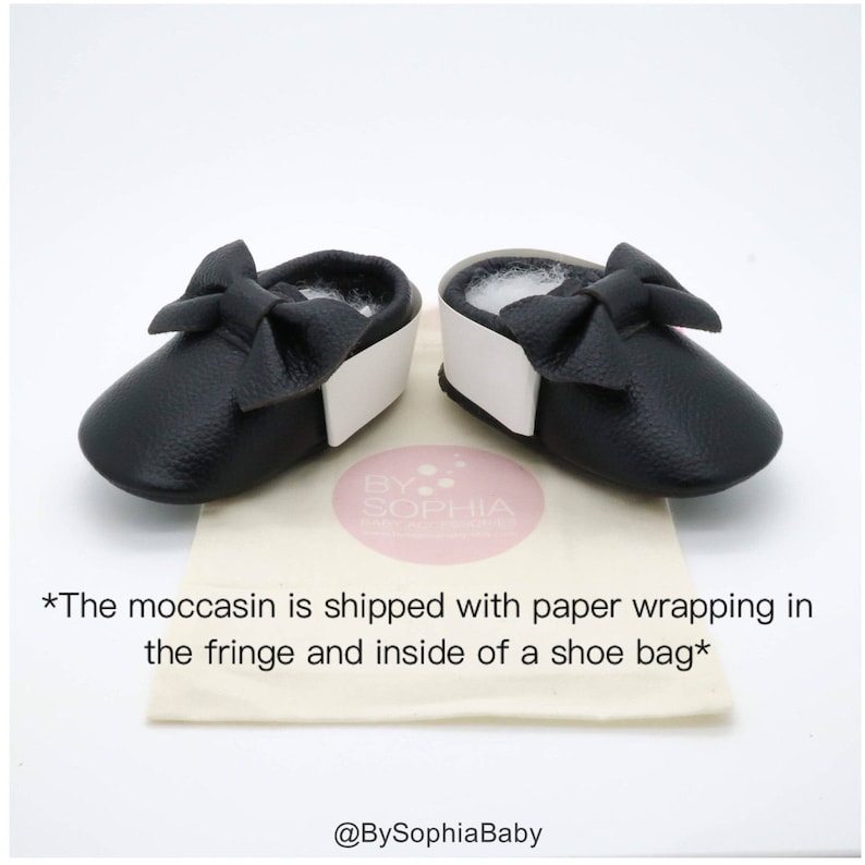 May include: A pair of black leather baby moccasins with a white trim and a black bow on each shoe. The moccasins are on a white and brown surface with the text "BY SOPHIA Baby Boutique" and "*The moccasin is shipped with paper wrapping in the fringe and inside of a shoe bag*" printed on the surface. The text "@BySophiaBaby" is also printed on the surface.