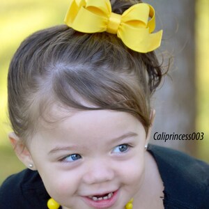 May include: A young girl with brown hair wearing a black shirt and a yellow beaded necklace. She is smiling and has a yellow bow in her hair. The background is blurred.