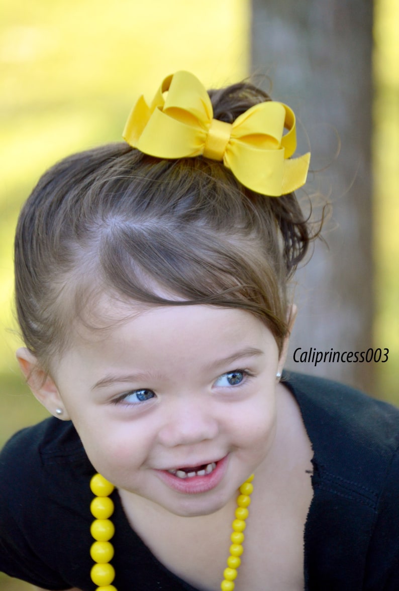 May include: A young girl with brown hair wearing a black shirt and a yellow beaded necklace. She is smiling and has a yellow bow in her hair. The background is blurred.