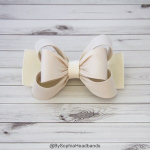 May include: A large, cream-colored satin bow with a white center. The bow is on a white wooden background.