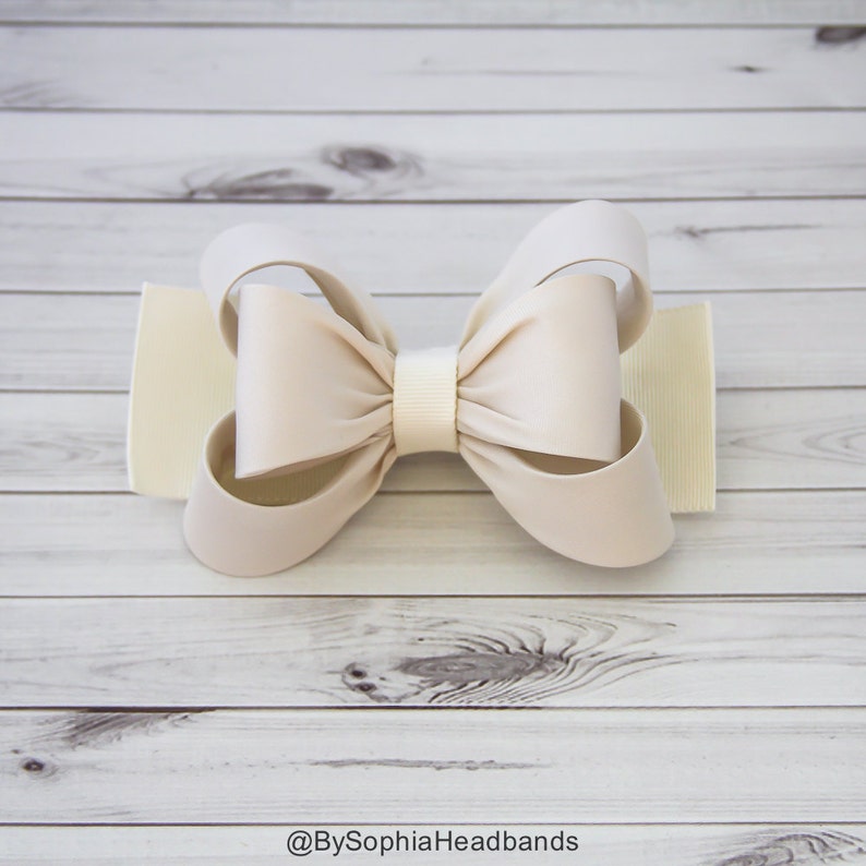 May include: A large, cream-colored satin bow with a white center. The bow is on a white wooden background.