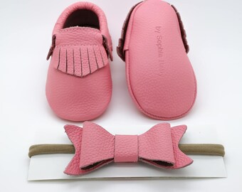 May include: Pink leather baby moccasins with fringe detail, a matching pink leather bow headband, and a brown elastic headband. The moccasins have the text "by Sophia Baby" on the side.