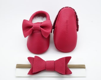 May include: A set of pink leather baby moccasins with a bow on the front, and a matching pink leather headband with a bow. The moccasins have the text "By Sophia" on the back.