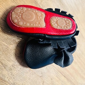 May include: Black leather baby moccasins with red trim and a heart-shaped sole. The moccasins have a fringe detail and a bow on the top.