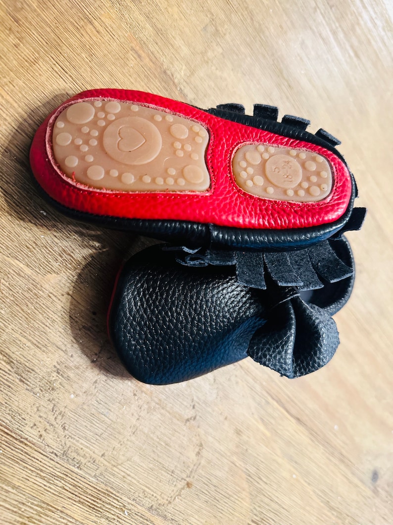 May include: Black leather baby moccasins with red trim and a heart-shaped sole. The moccasins have a fringe detail and a bow on the top.