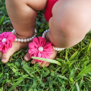 Pode incluir: Um par de sand&aacute;lias de beb&ecirc; de flores rosa com detalhes de p&eacute;rolas brancas. As sand&aacute;lias s&atilde;o usadas por um beb&ecirc; em um leito de grama verde.