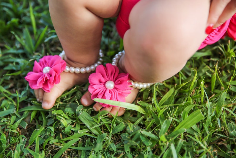 Pode incluir: Um par de sand&aacute;lias de beb&ecirc; de flores rosa com detalhes de p&eacute;rolas brancas. As sand&aacute;lias s&atilde;o usadas por um beb&ecirc; em um leito de grama verde.