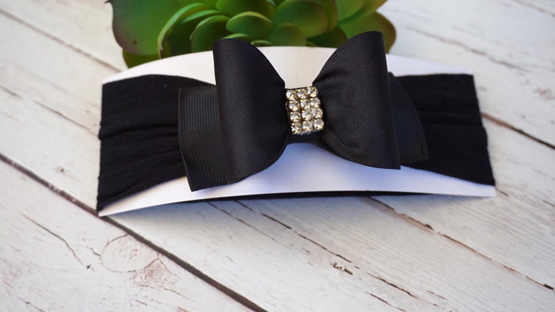 May include: A black baby headband with a large bow and a rhinestone embellishment. The bow is centered on the headband and is made of a smooth, shiny fabric. The headband is made of a soft, stretchy material.