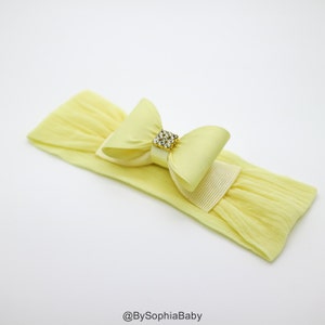 May include: A yellow headband with a yellow satin bow and a gold rhinestone embellishment. The headband is made of stretchy fabric.
