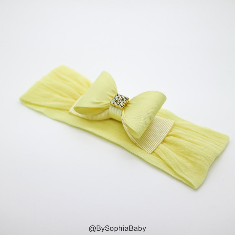 May include: A yellow headband with a yellow satin bow and a gold rhinestone embellishment. The headband is made of stretchy fabric.