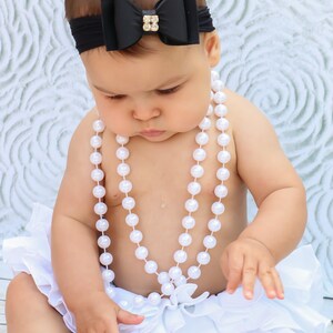 May include: A baby wearing a black bow headband with a gold gem center, a white ruffled diaper cover, and a pearl necklace. The baby is sitting on a wooden surface.